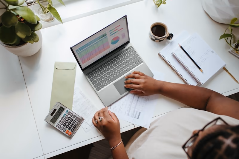 Seen from above, a person at a white desk with a laptop of figures, a calculator and a printed invoice