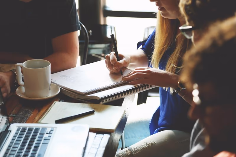 Three people around a cafe table, one writing in an open notebook