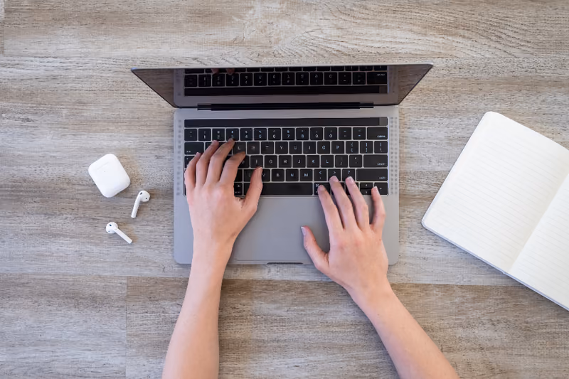 Hands typing on a laptop on a wooden desk, a closed notebook beside it