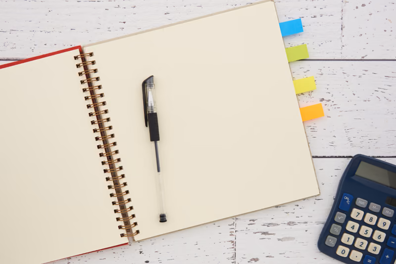 An open notebook with coloured page tabs beside a calculator, on whitewashed wood