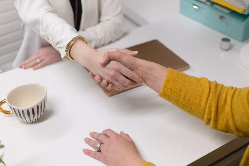Two people shaking hands over a desk, one in a mustard-yellow sleeve