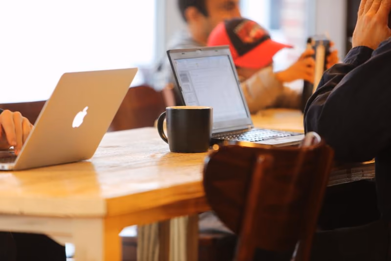 Two open laptops and a mug on a wooden cafe table, with other people working behind