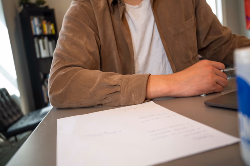 A person at a desk reading a printed page with a pen in hand