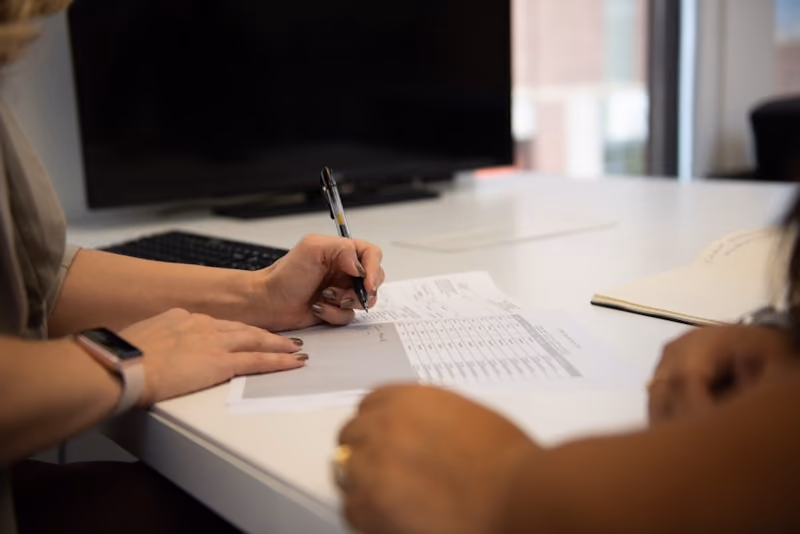 Two people at a desk, one filling in a printed form with a pen