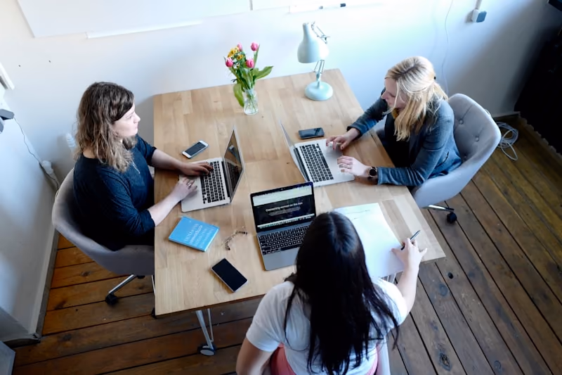 Three people working around a wooden table, seen from above, each with a laptop or an open notebook