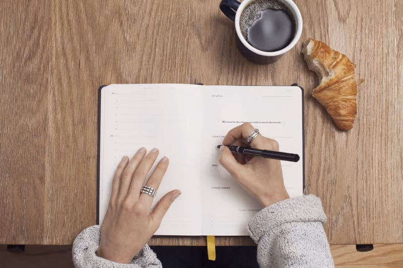 Hands writing in an open notebook on a wooden table, beside a coffee and a croissant