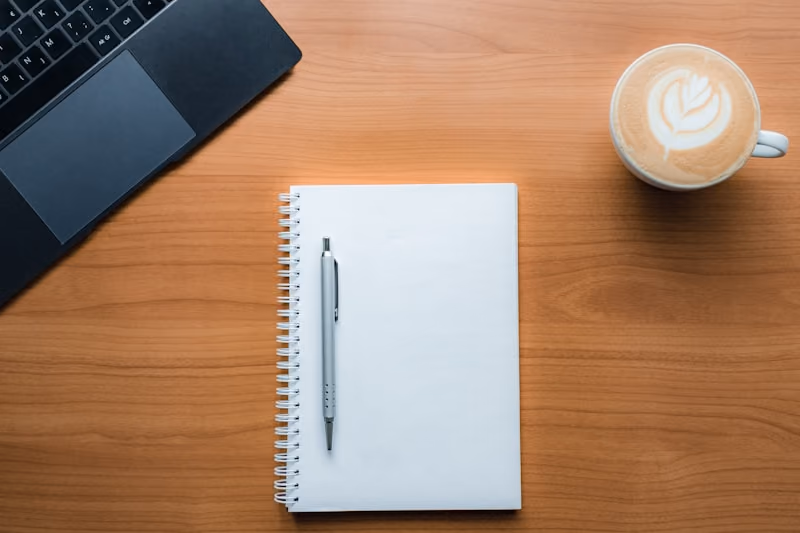 An open notebook and a latte on a wooden table, beside the corner of a laptop